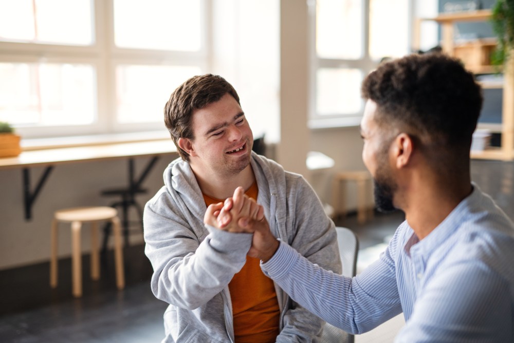 Young happy man with Down syndrome with his mentoring friend celebrating success indoors at school